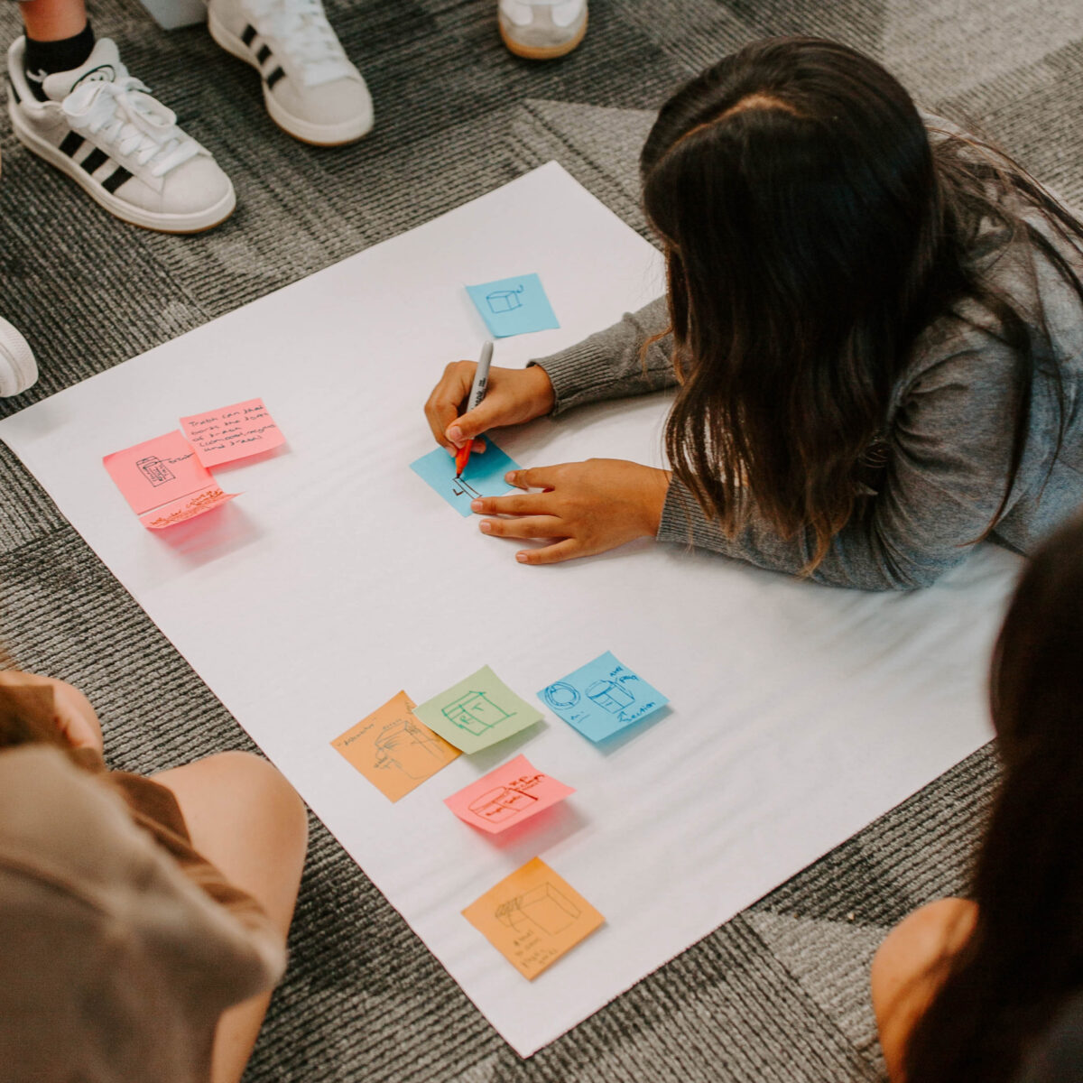 Young student sketching an idea on a sticky note while sitting on the floor with others during a collaborative workshop.